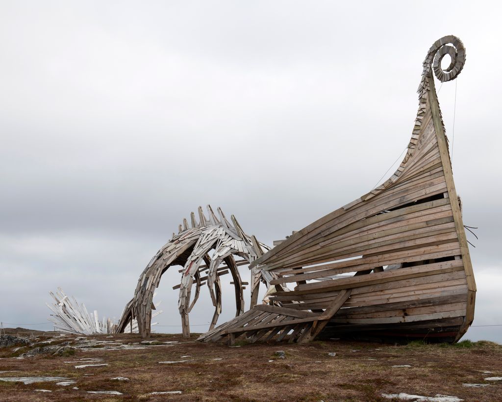 Treskulptur ute i et viddelandskap. Formen minner om baugen på et vikingskip som går over i et dyreskjelett og ender opp i en stjert. Foto.