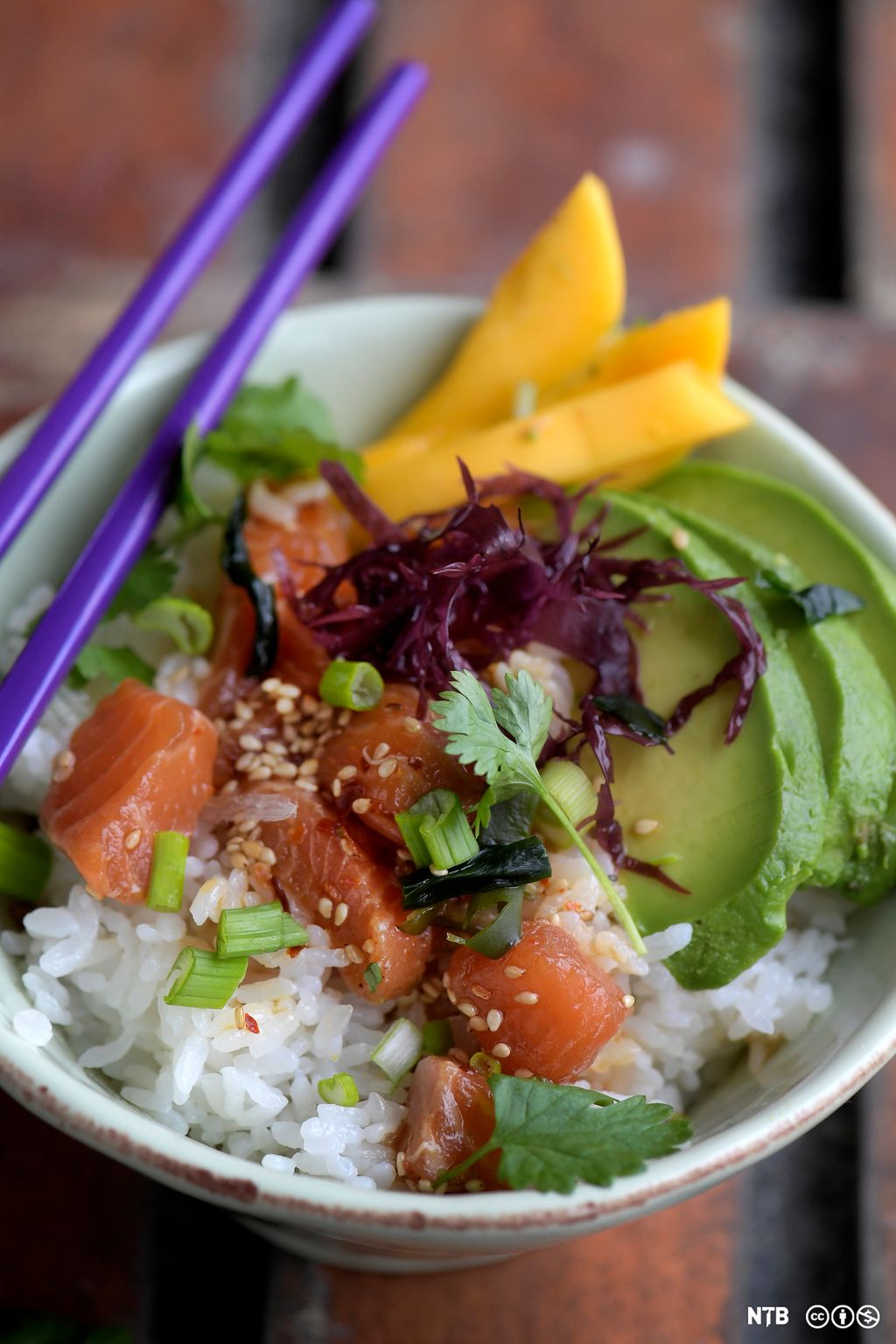 En bolle med rå laks i terninger, hvit ris, avokado i skiver, rødkål, mango og vårløk. Foto.