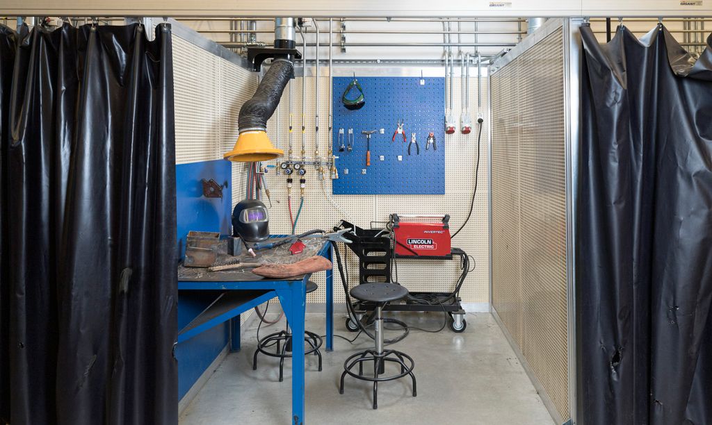 Sveisebås med maskiner og utstyr. Arbeidsbenk med vernesutstyr, avtrekk og lystett forheng. Foto.