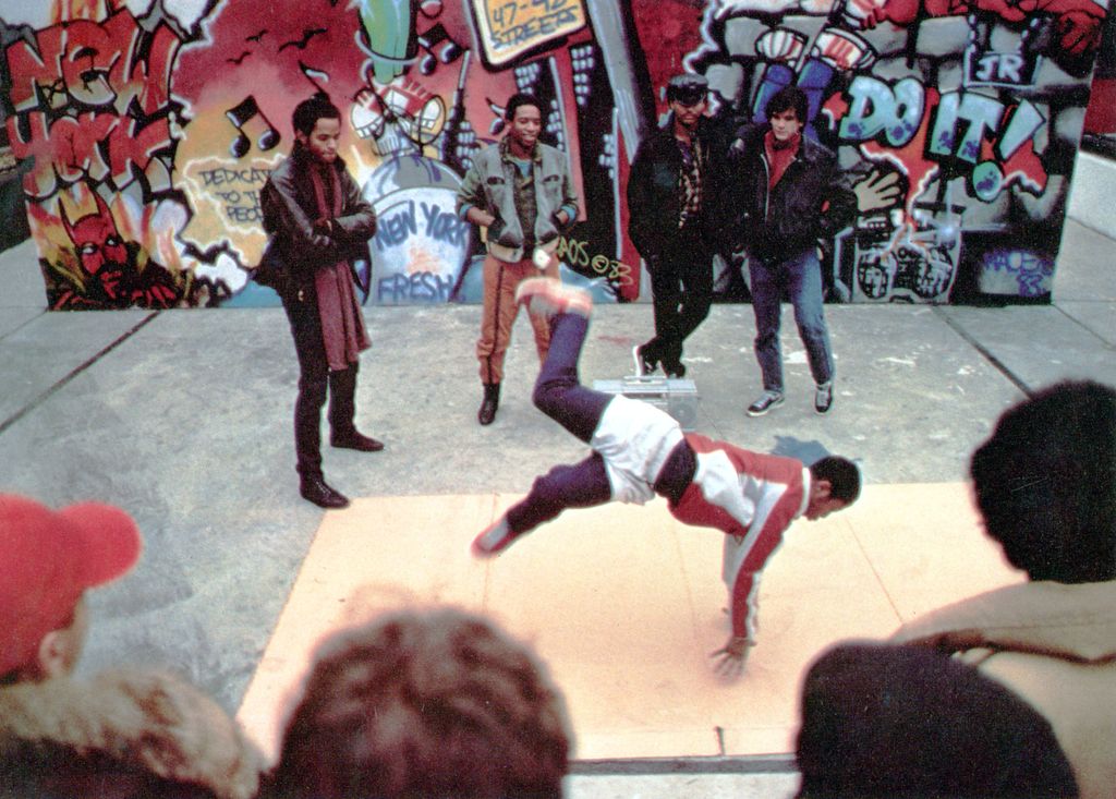 En person driver break dancing på gata på en utbrettet pappkartong. Flere personer ser på. I bakgrunnen er det en vegg med graffiti på. Foto.
