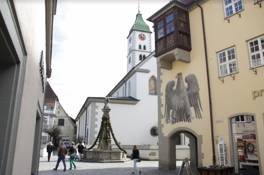 Wangen i Tyskland: Et torg med en brønn som er pynta med grangirlander. Ved torget ei kirke og en bygning med et stort våpenskjold og et gammelt karnappvindu på veggen. Foto.