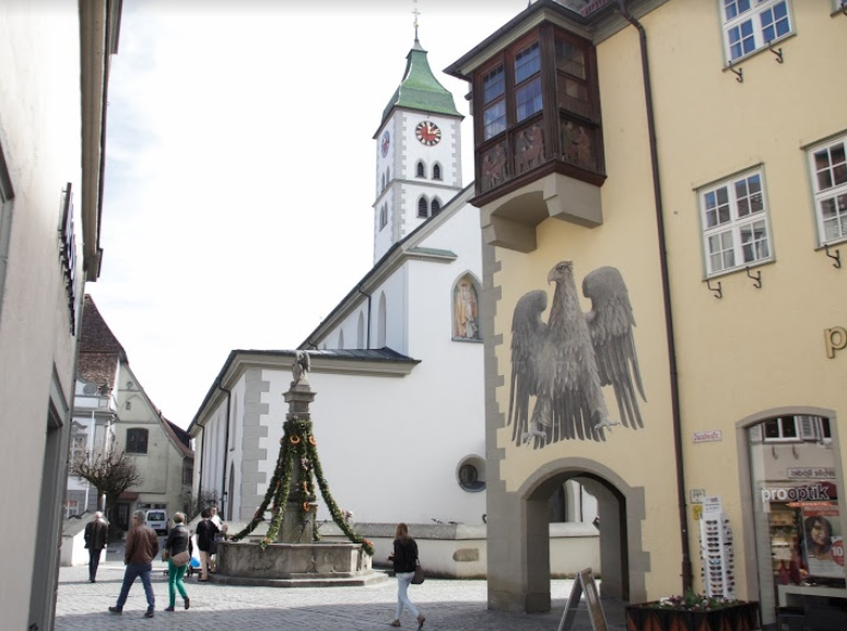 Wangen i Tyskland: Et torg med en brønn som er pynta med grangirlander. Ved torget ei kirke og en bygning med et stort våpenskjold og et gammelt karnappvindu på veggen. Foto.