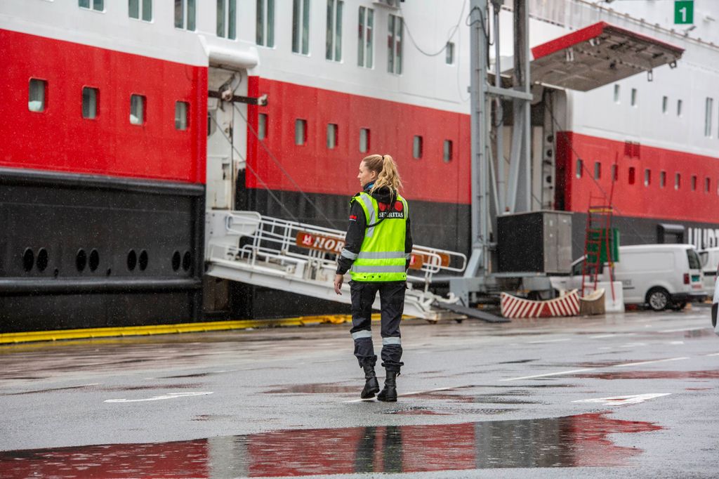 Uniformert vekter står på en kai med ryggen til kameraet, vendt mot et hurtigruteskip som er lagt til kai. Foto.