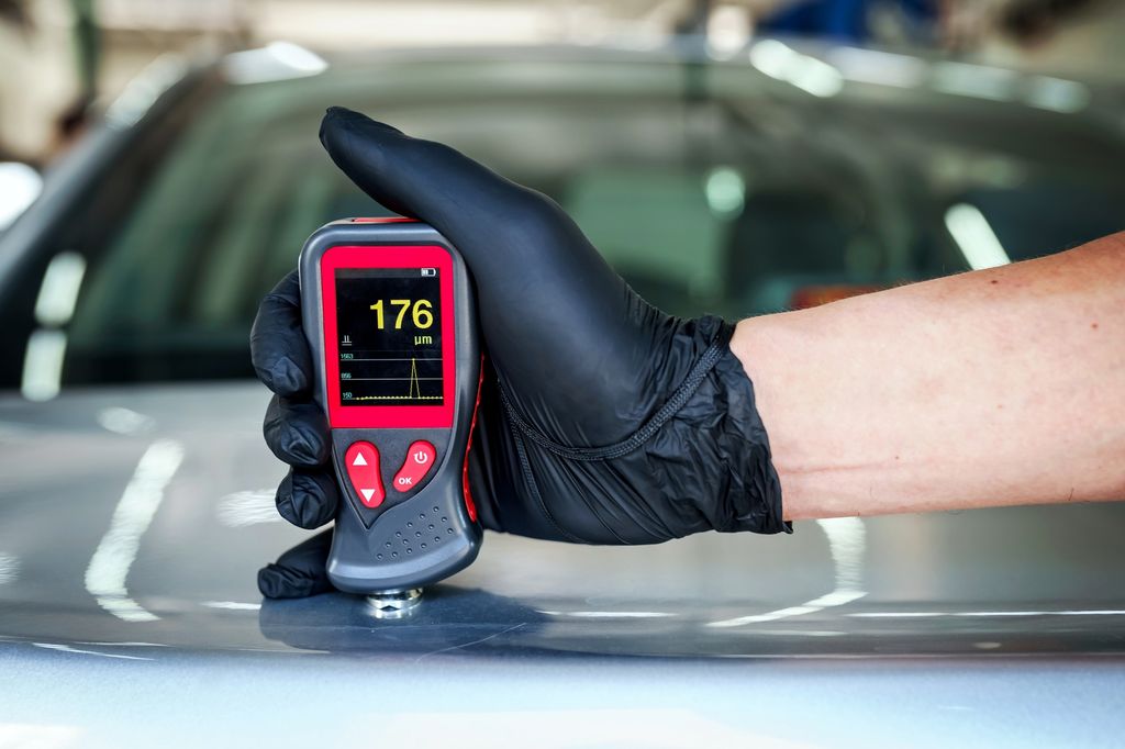 Hånd som holder et måleapparat for måling av lakktykkelse mot ei lakkert overflate. Displayet viser 176 μm. Foto.