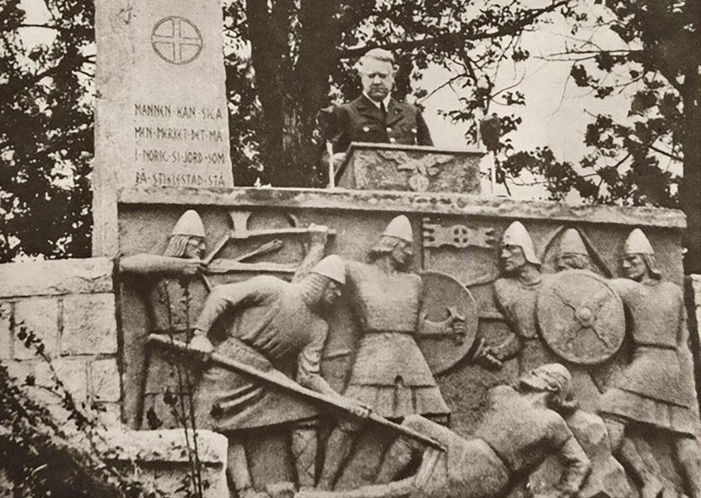 Quisling står på en talerstol bak monumentet som er et stort steinrelieff. Foto.