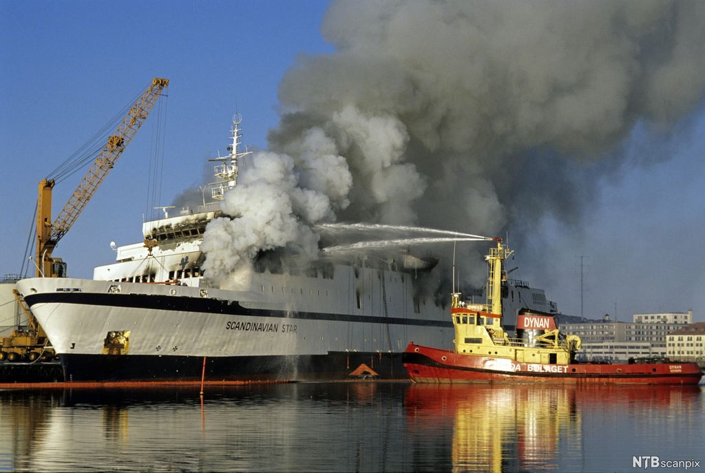 Tjukk, grå røyk velter ut av en stor passasjerferge. Ved siden av fergen ligger en brannbåt som driver slokningsarbeid. Foto.