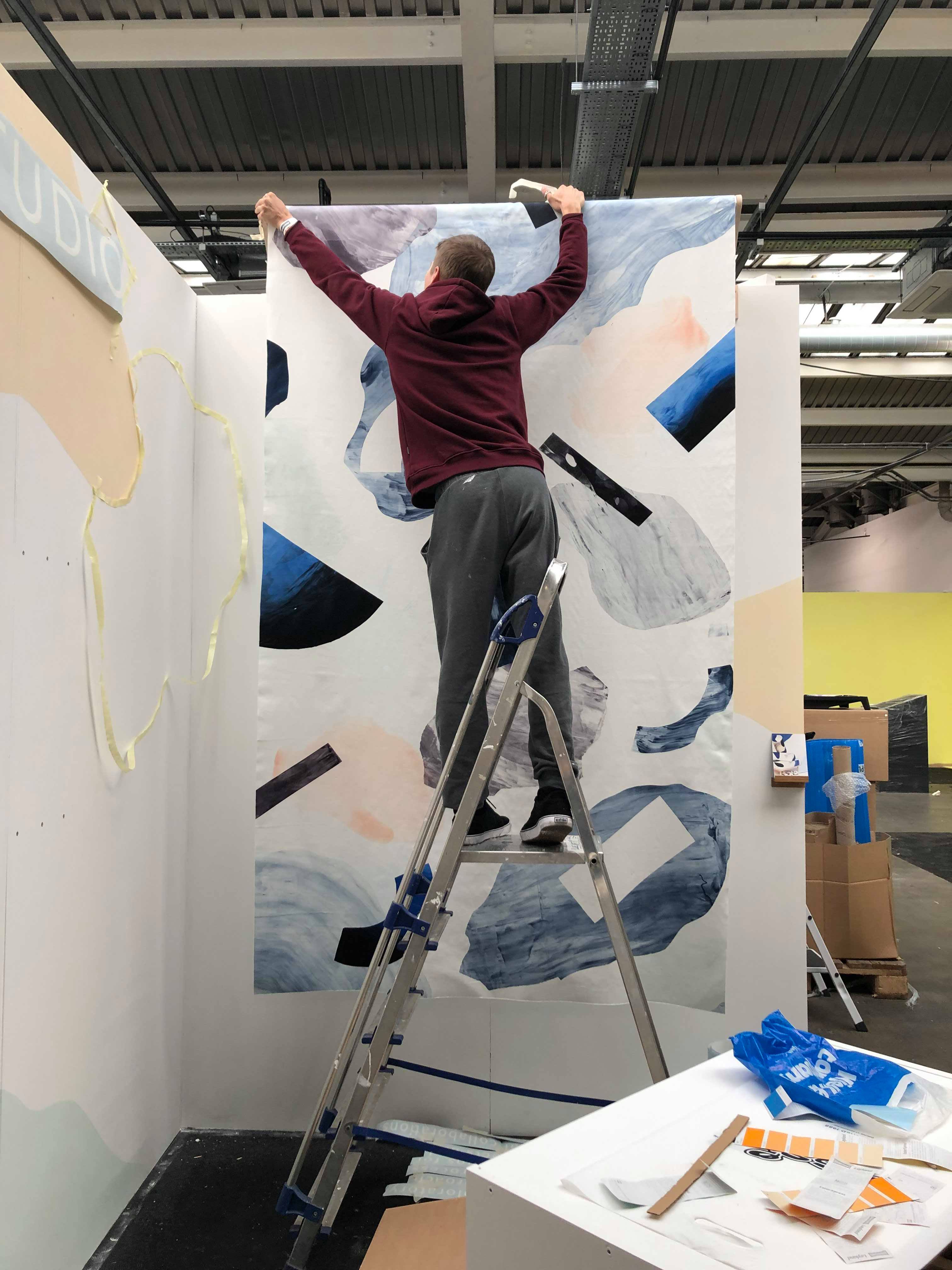 Fotografi av mann som står i ei gardintrapp og henger opp et tekstiltrykk.