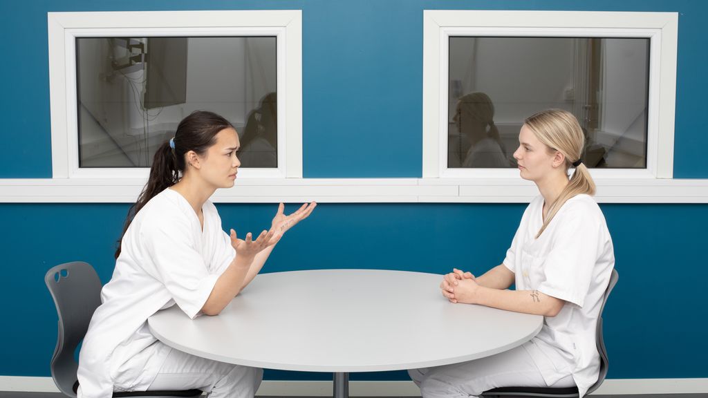 To kvinner i uniform for helsepersonell sit ved eit rundt bord inntil ein vegg. Den eine kvinna uttrykker seg tydeleg med hendene og har eit alvorleg og litt frustrert ansiktsuttrykk. Den andre kvinna sit med hendene samla og lyttar roleg til det som blir sagt. Foto.
