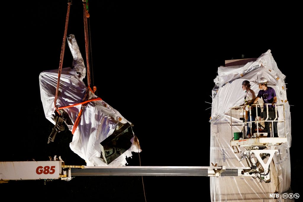 Sculpture wrapped in plastic is hoisted in the air by a crane. Two men are on the crane. Photo.