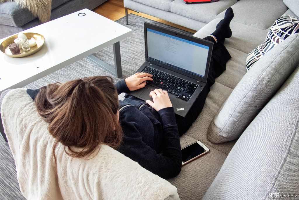 Jente ligger på sofa med PC og mobil. Foto.