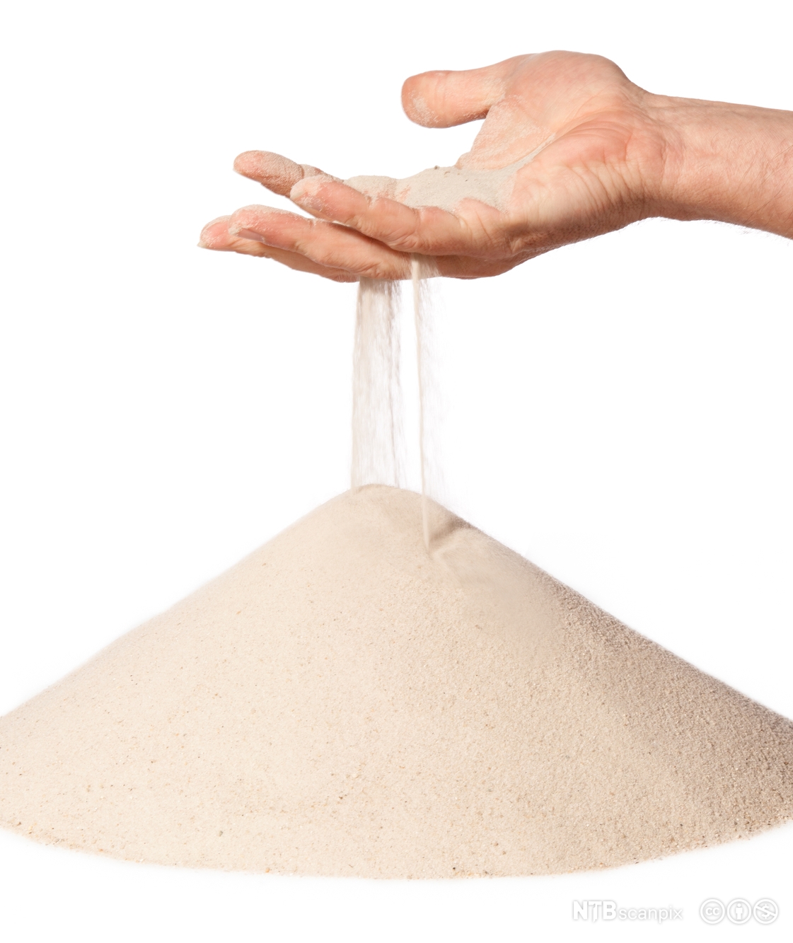 Fin sand renner mellom fingrene på ei hånd og danner en haug mot en hvit bakgrunn. Foto.
