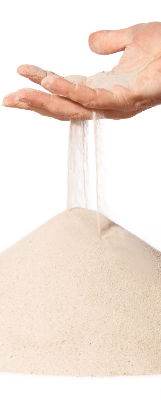 Fin sand renner mellom fingrene på ei hånd og danner en haug mot en hvit bakgrunn. Foto.