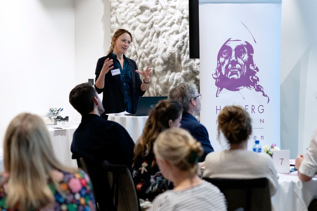 Kvinne held foredrag for ei forsamling. Ved sida av henne står eit banner med teksten "Holbergprisen i skolen". Foto.