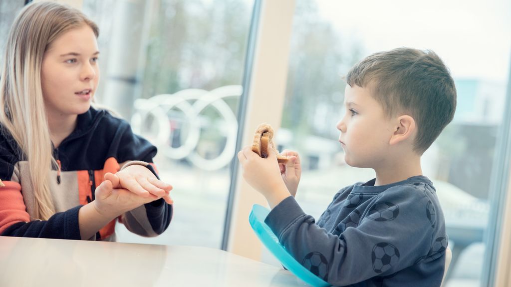 Voksen leder og liten gutt i blå fotballgenser snakker sammen under spisetid på SFO. Den voksne ser på gutten og holder håndflatene mot hverandre med hendene flatt litt over bordet. Gutten river i brødskiva si. Foto.
