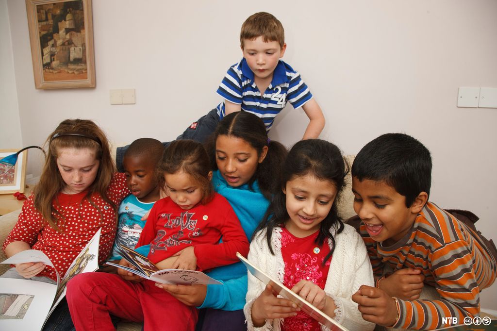 Seks barn sitter på rekke i en sofa og ser i bøker. En gutt sitter litt høyere bak dem og ser også i den ene boka. Foto.