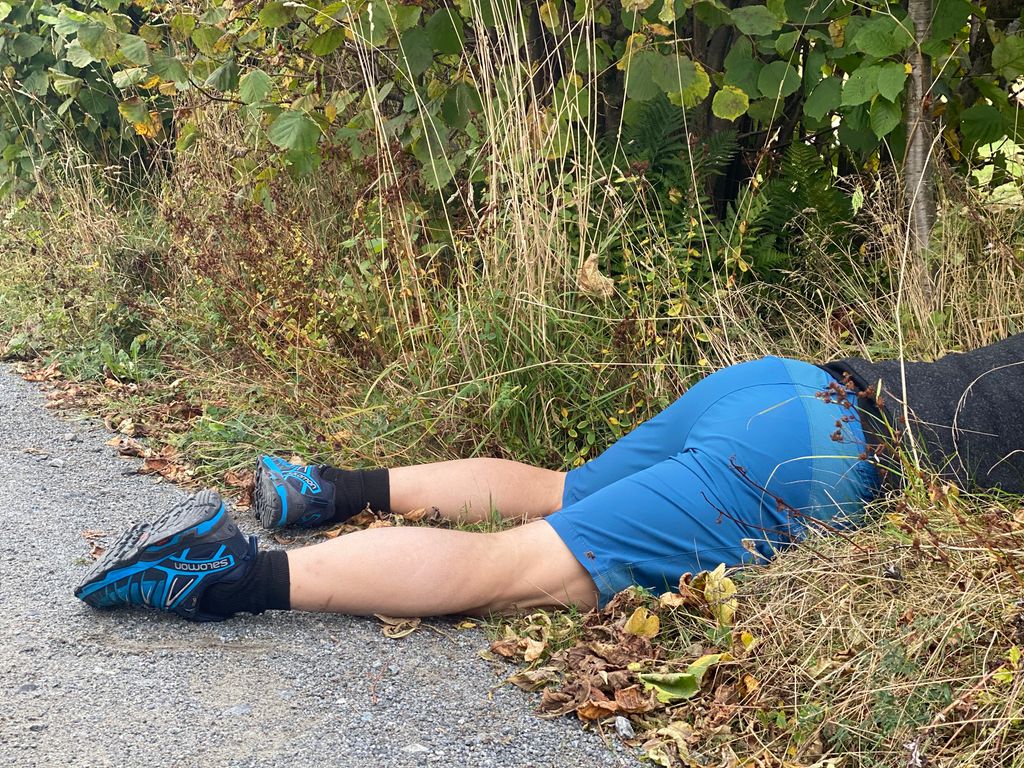 En person ligger på magen ved siden av veien med føttene i veibanen. Vi ser bare underkroppen og beina. Personen har joggesko og treningstøy. Foto. 