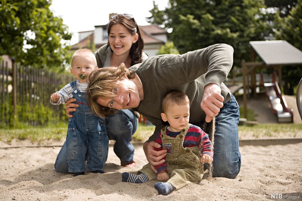 To små barn og to voksne i ei sandkasse på en lekeplass. Det ene barnet blir holdt stående av en kvinne, og det andre barnet sitter i sanda mens en mann støtter. Mannen lar sand gli ut av hånda si, og det lille barnet ser på sanda. Foto. 