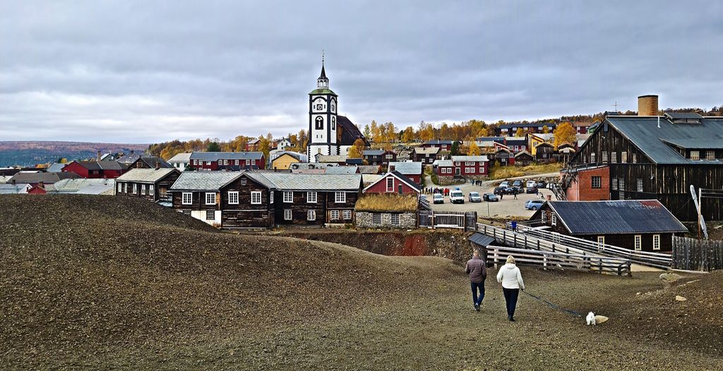 Slagghaugene med slagget fra gruvevirksomheten på Røros i forkant. To personer går tur med en liten hund. Eldre trebygninger og Røros kirke i bakgrunnen. Foto. 