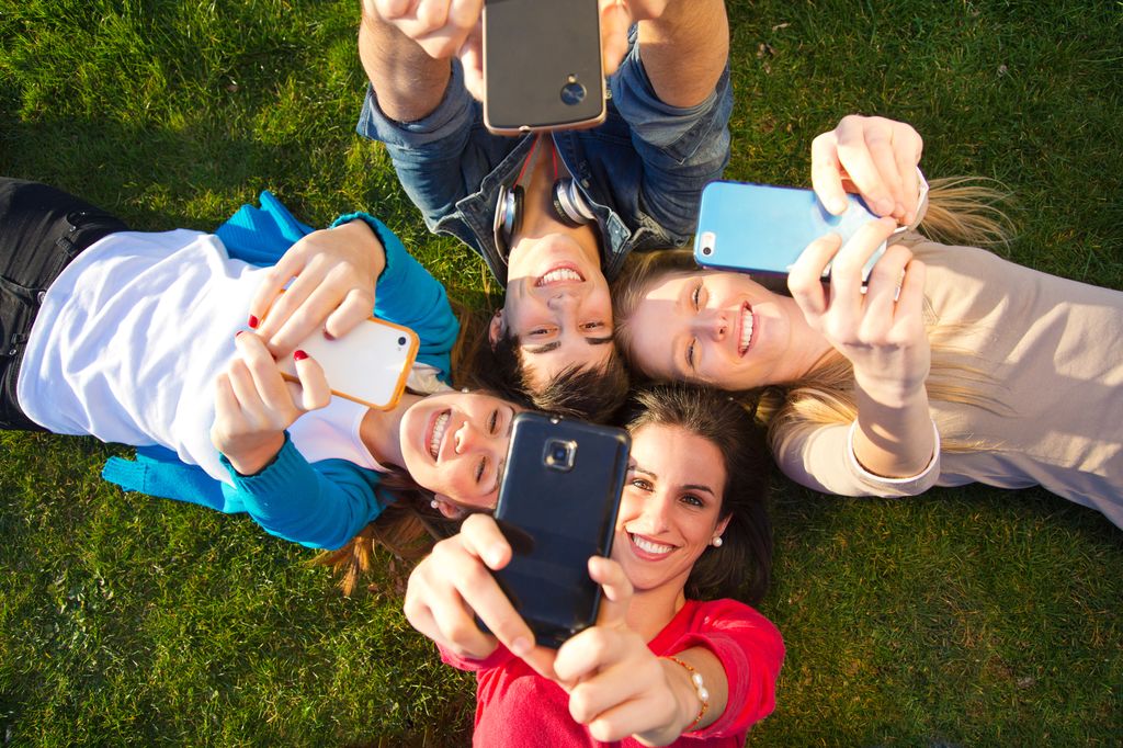 Fire smilande ungdommar ligg på ei mark og tek ein selfie. Foto.
