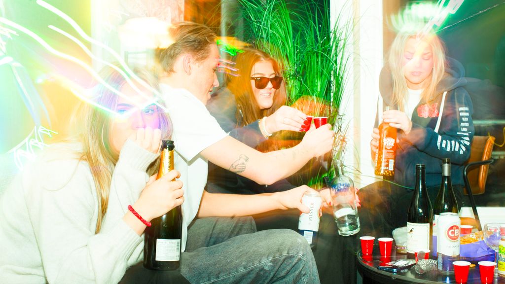 Fire ungdommer er samlet rundt et bord. Det er flasker med alkohol på bordet, og to av ungdommene skåler med hverandre. Ei av jentene kikker bort fra de andre og ser usikker ut. Foto.