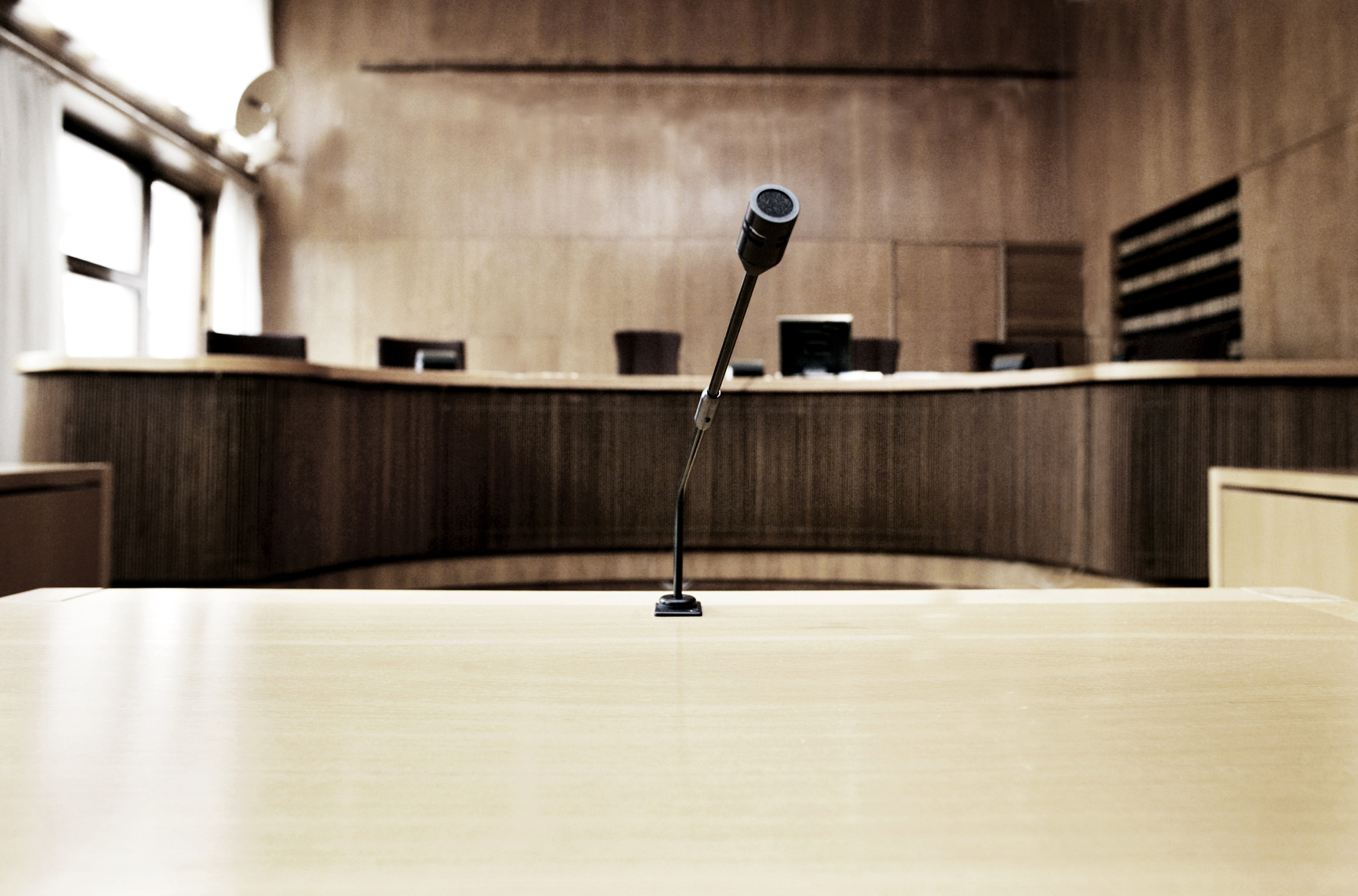 Fem tomme stoler er plassert bak en skranke på et litt opphøyd podium. I forgrunnen er det et mikrofonstativ plassert på et bord. Foto.