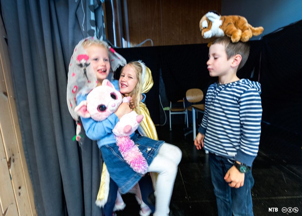 Elever som leker teater. Ei jente med gullkappe med hette over hodet bærer ei jente med et tøystykke på hodet. Jenta som blir båret, holder en stor, rosa bamse. En gutt står ved siden av og balanserer en liten tøytiger på hodet. Foto.