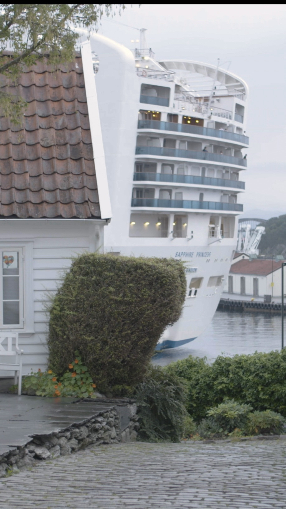 Eit svært cruiseskip dukkar opp bak eit kvitmåla trehus. Foto.