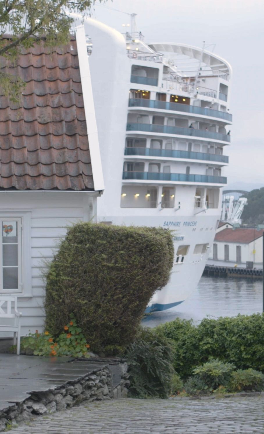 Eit svært cruiseskip dukkar opp bak eit kvitmåla trehus. Foto.