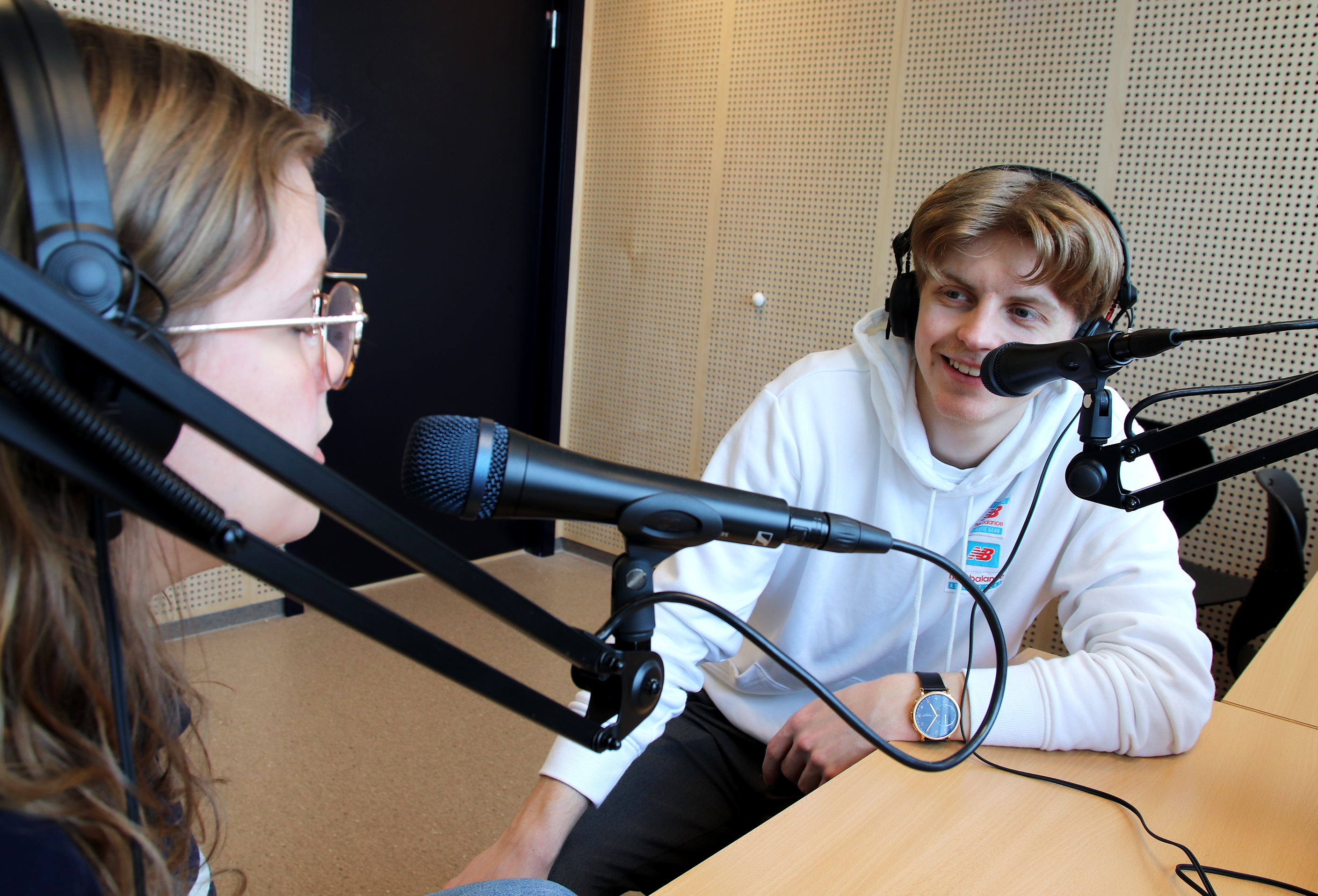 To elever i samtale i lydstudio. De har på øreklokker og sitter foran hver sin mikrofon. Foto.