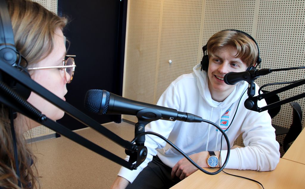 To elever i samtale i lydstudio. De har på øreklokker og sitter foran hver sin mikrofon. Foto.