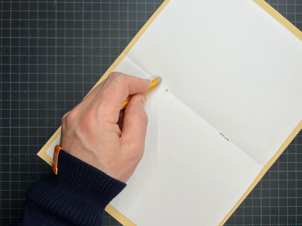 Ei hånd bruker en blyant til å bøye en stift i bretten på et hefte med ark. Foto.