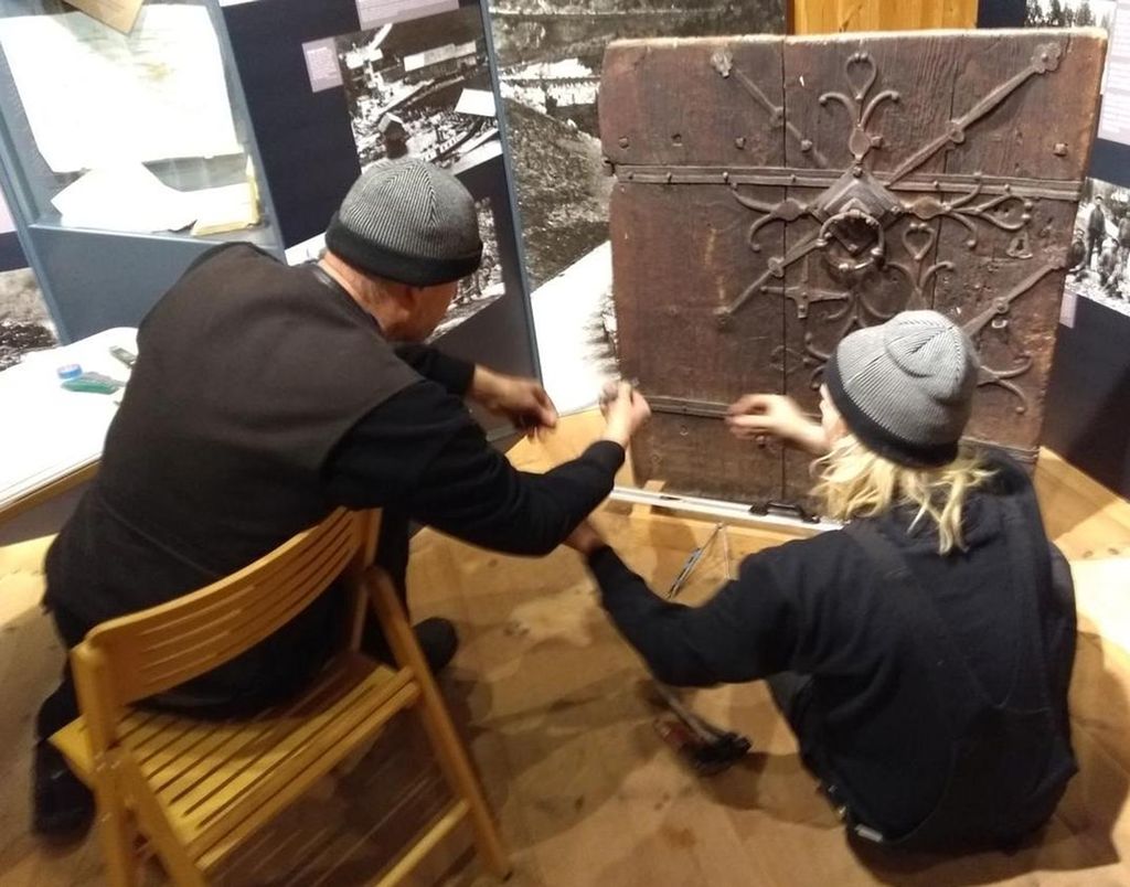 To menn samarbeider om å ta mål av et jernbeslag på ei dør. Foto.