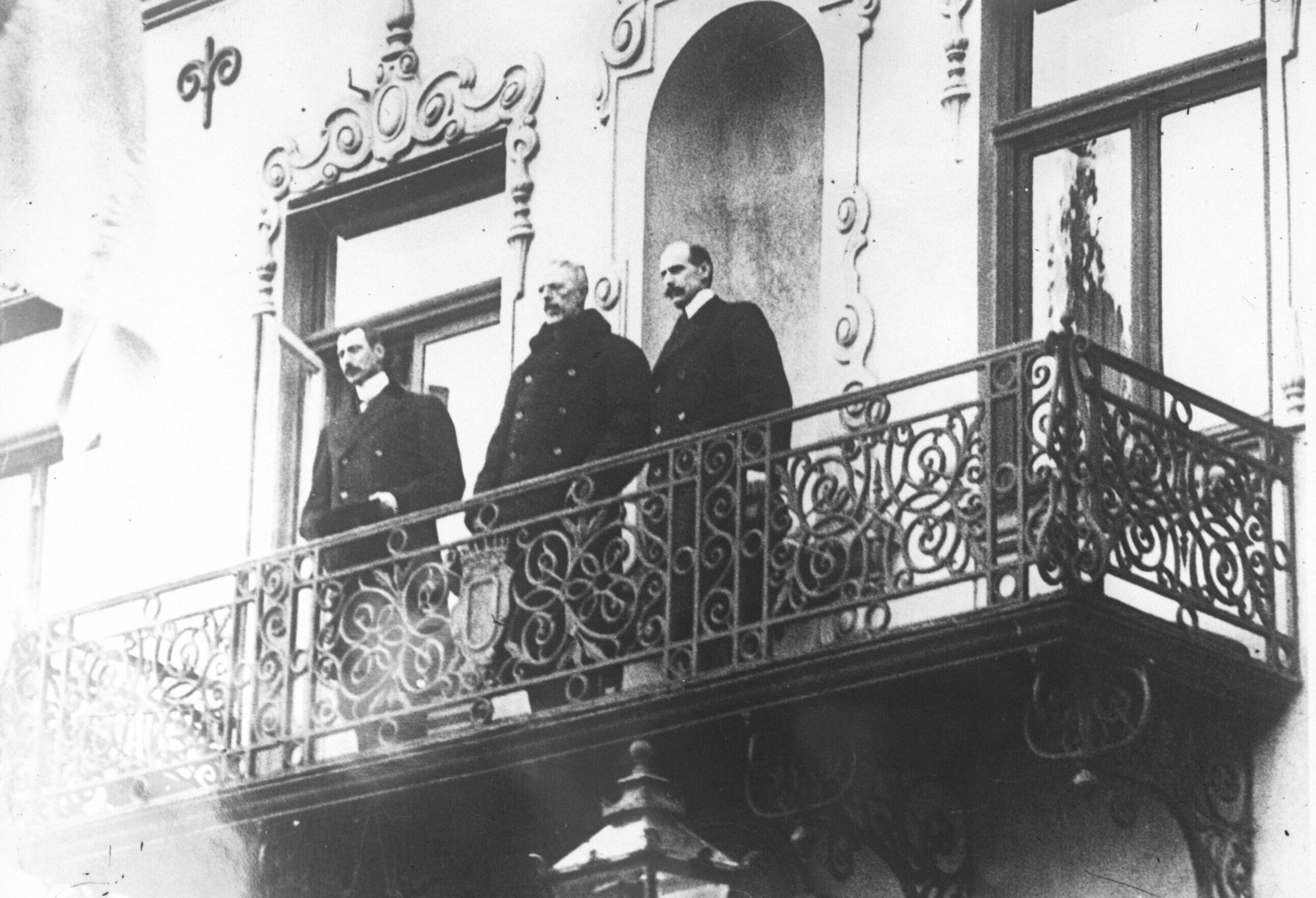 De tre kongene, Haakon 7. av Norge, Gustav 5. av Sverige og Christian 10. av Danmark, står i vinterfrakker og med hattene av oppe på en smijernsbalkong. Foto. 