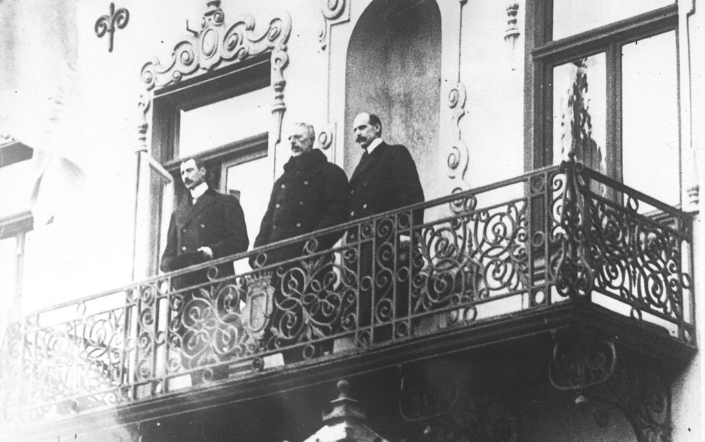 De tre kongene, Haakon 7. av Norge, Gustav 5. av Sverige og Christian 10. av Danmark, står i vinterfrakker og med hattene av oppe på en smijernsbalkong. Foto.