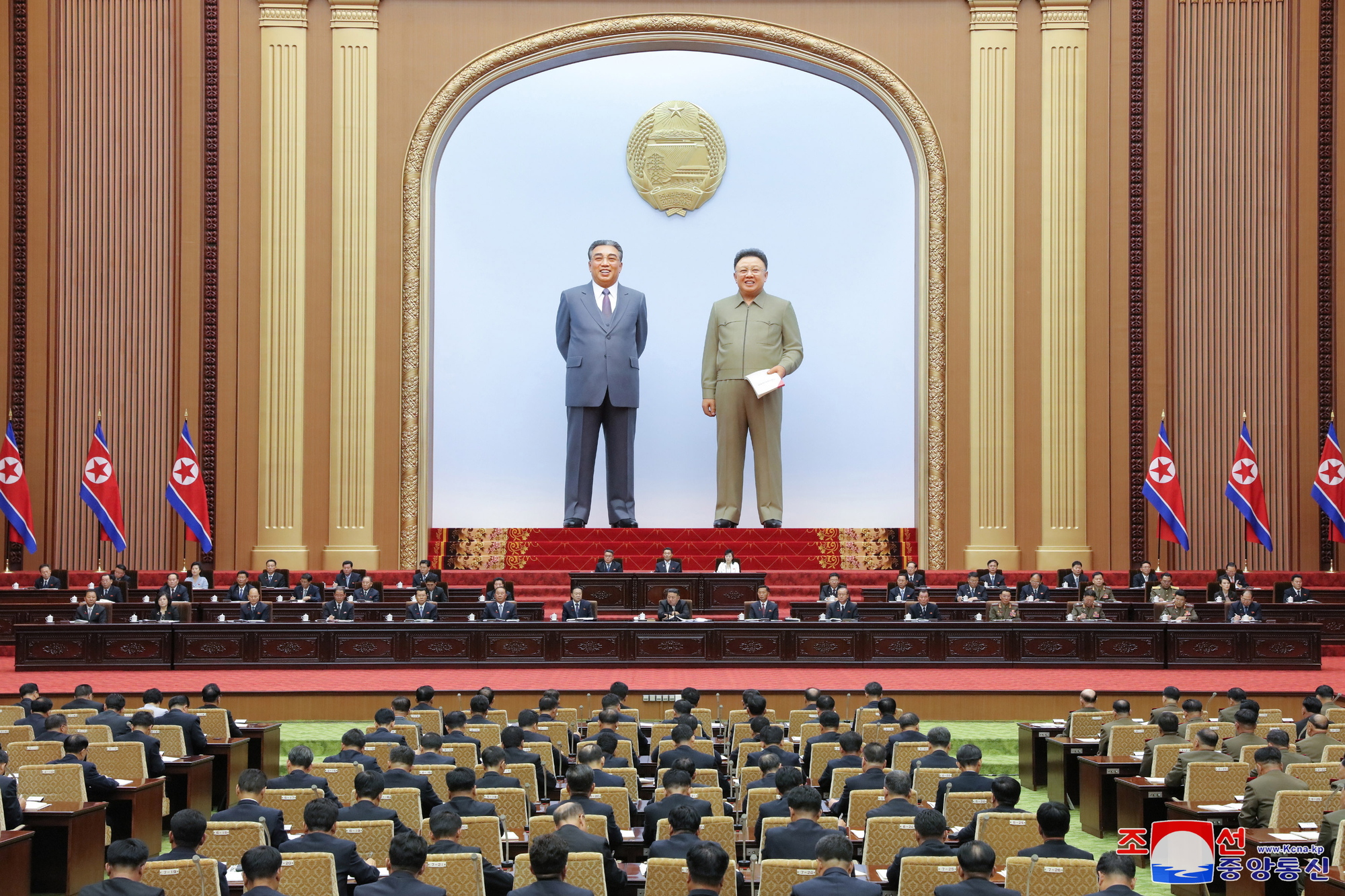 To enorme, naturtru  statuar står på ein scene bakarst i ein sal. Framfor dei sit  to-tre rekker på eit opphøgd podium. I salen, vende mot dei, sit mange på rekke og rad. Nordkoreanske flagg står på kvar side. Foto.