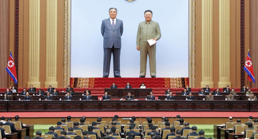 To enorme, naturtru statuar står på ein scene bakarst i ein sal. Framfor dei sit to-tre rekker på eit opphøgd podium. I salen, vende mot dei, sit mange på rekke og rad. Nordkoreanske flagg står på kvar side. Foto.