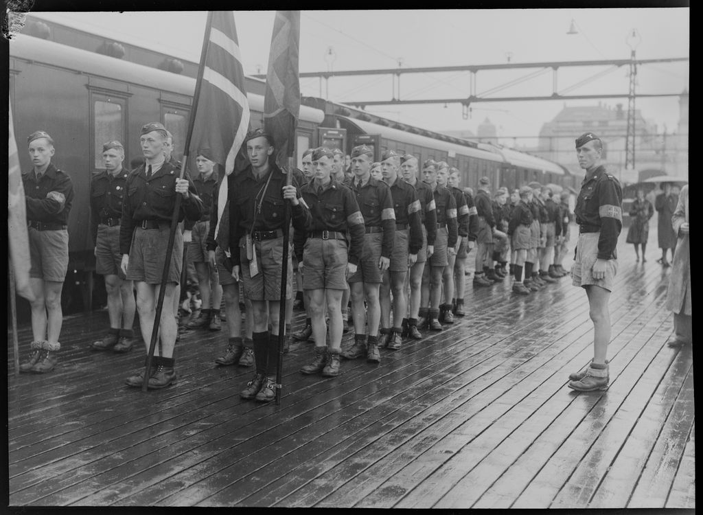 Unggutter i hirduniform står militært oppstilt i regnvær på perrongen ved siden av et tog. I bakgrunnen står det et par kvinner under paraplyer. Foto.