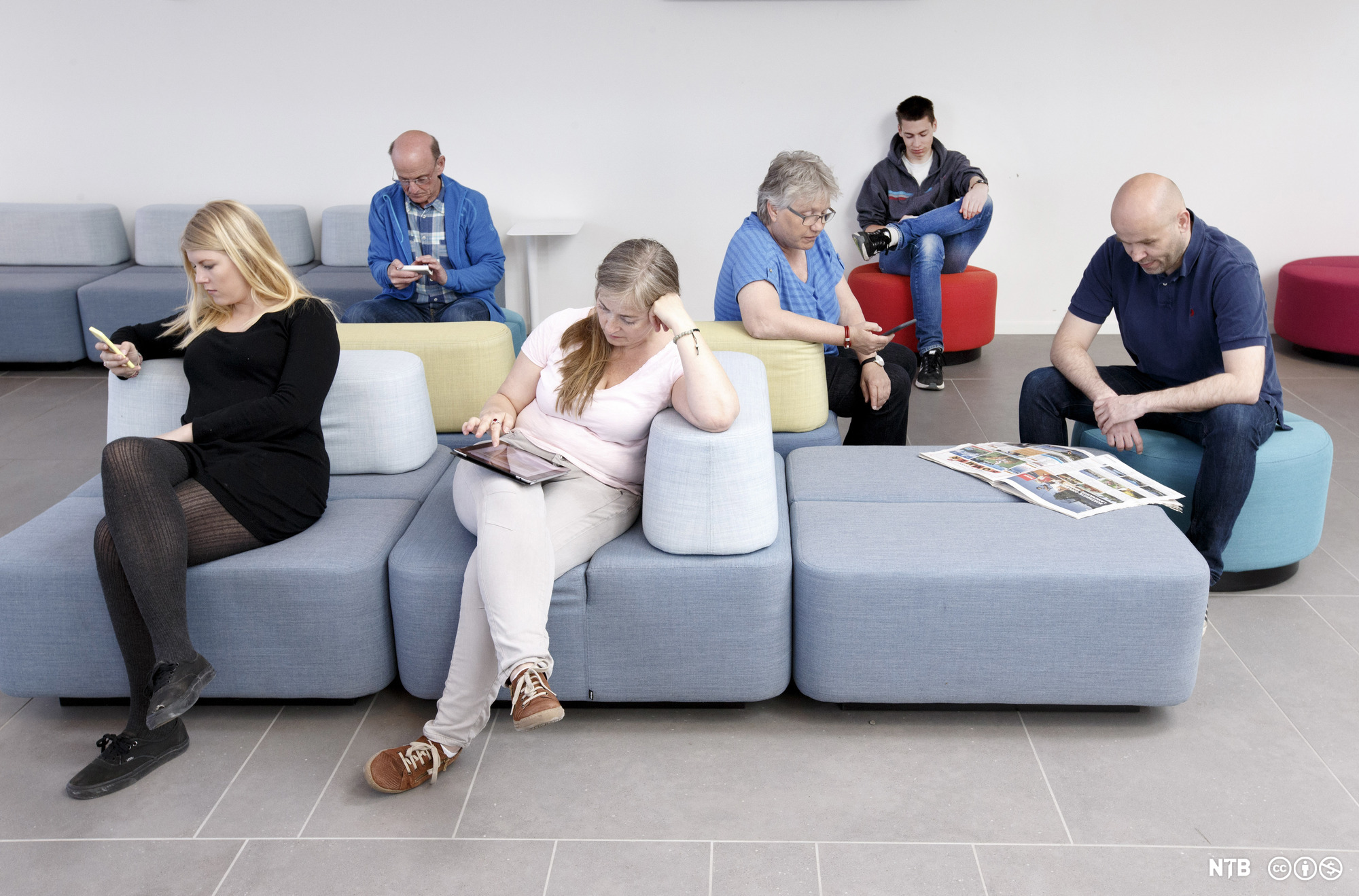 Kvinner og menn i ulike aldrar sit i sofaer og på puffar. Kvar av dei ser på anten ei avis, eit nettbrett eller ein smarttelefon. Foto.