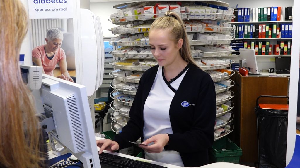 Apotektekniker står ved en pc og sjekker ID-kortet til en kunde. Foto.