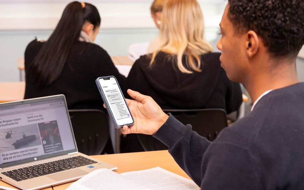 Gutt sitter i et klasserom og bruker praterobot på mobil. På pulten foran ham er det ei oppslått bok og en bærbar datamaskin med ei nettavis på skjermen. Foto.