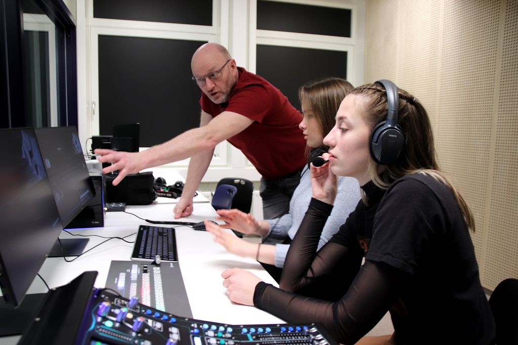 En lærer og to elever henholdsvis står og sitter foran flere skjermer, tastaturer og en bildemikser. Foto.