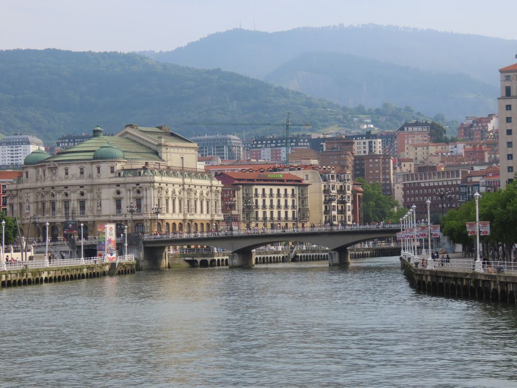 En elv med ei bru og flere store byninger i bakgrunnen. Foto.