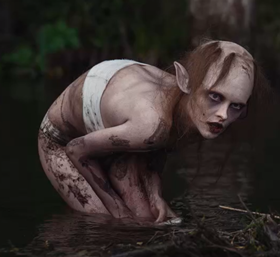 Ein nesten naken menneskeliknande figur står samankrøkt til knes i vatnet og har blikket festa på noko til høgre i biletet. Foto.