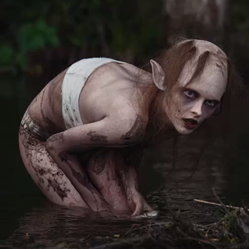 Ein nesten naken menneskeliknande figur står samankrøkt til knes i vatnet og har blikket festa på noko til høgre i biletet. Foto.