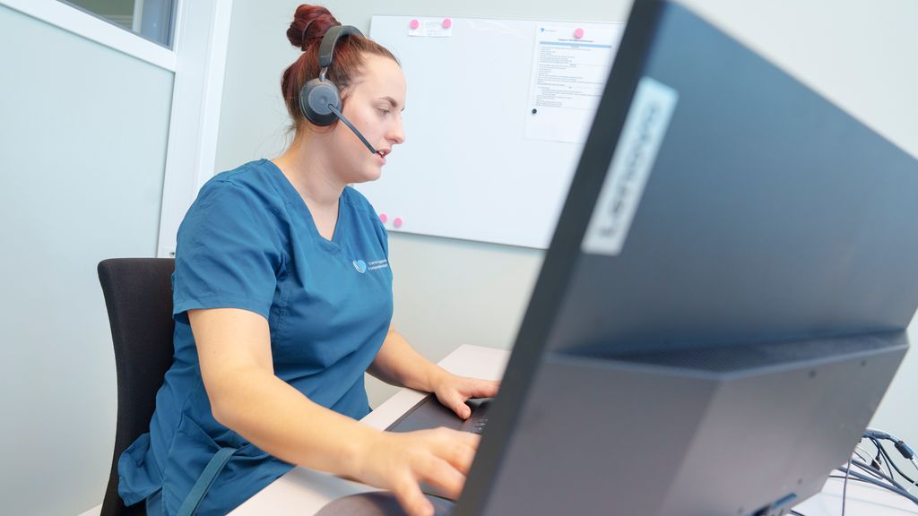 Helsesekretær med hodetelefoner med mikrofon sitter foran pc og skriver på tastaturet. Foto.