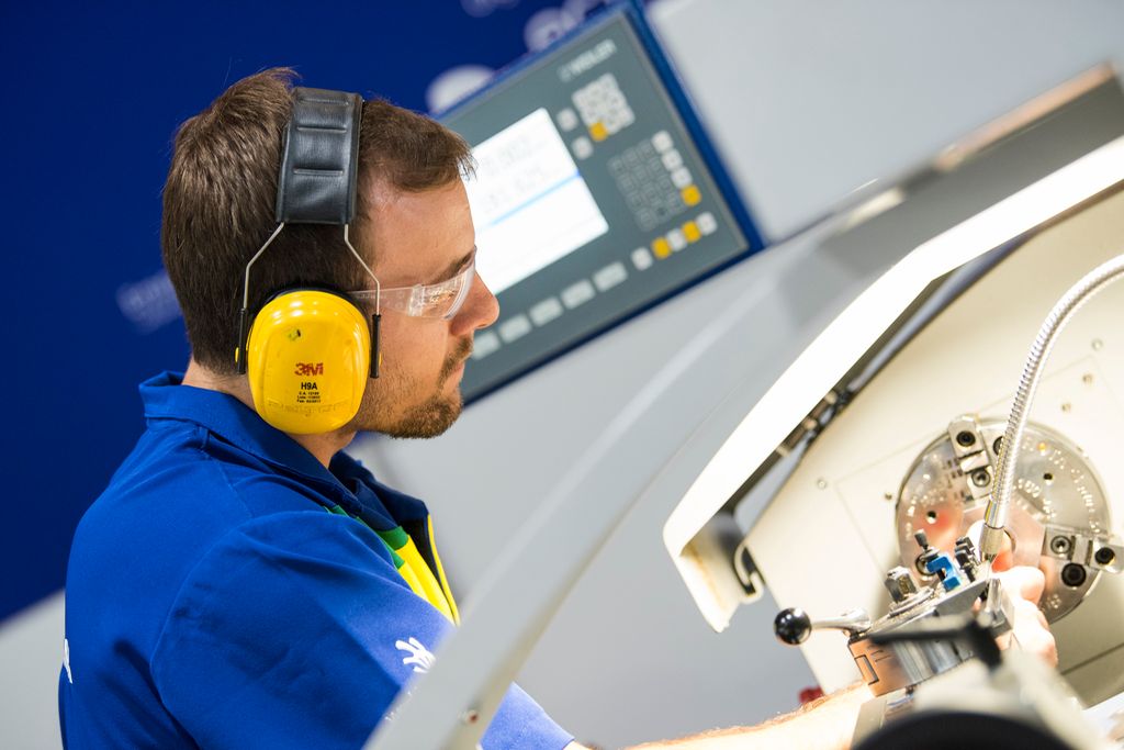 Person som står ved en dreiebenk. Han er ikledd arbeidsklær, hørselvern og vernebriller. Foto.