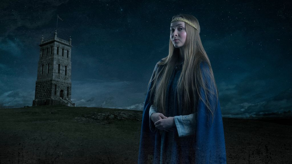 En ung kvinne med langt hår og blå drakt og kappe foran et tårn på et fjell nattestid. Manipulert foto.