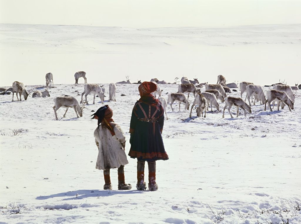 En samisk kvinne med kofte og et barn med kofte og pesk i forgrunnen, reinsdyr på snødekt vidde i bakgrunnen. Foto. 