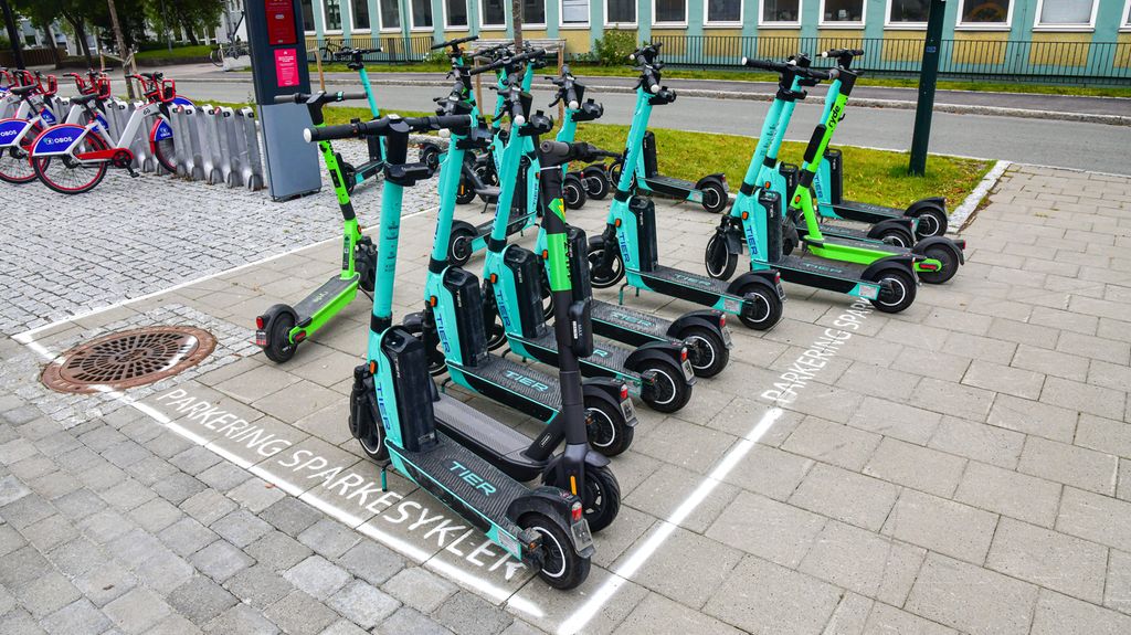 Elsparkesykler fra to ulike utleiefirmaer står på en parkeringsplass for sparkesykler. Bysykler i bakgrunnen. Foto.