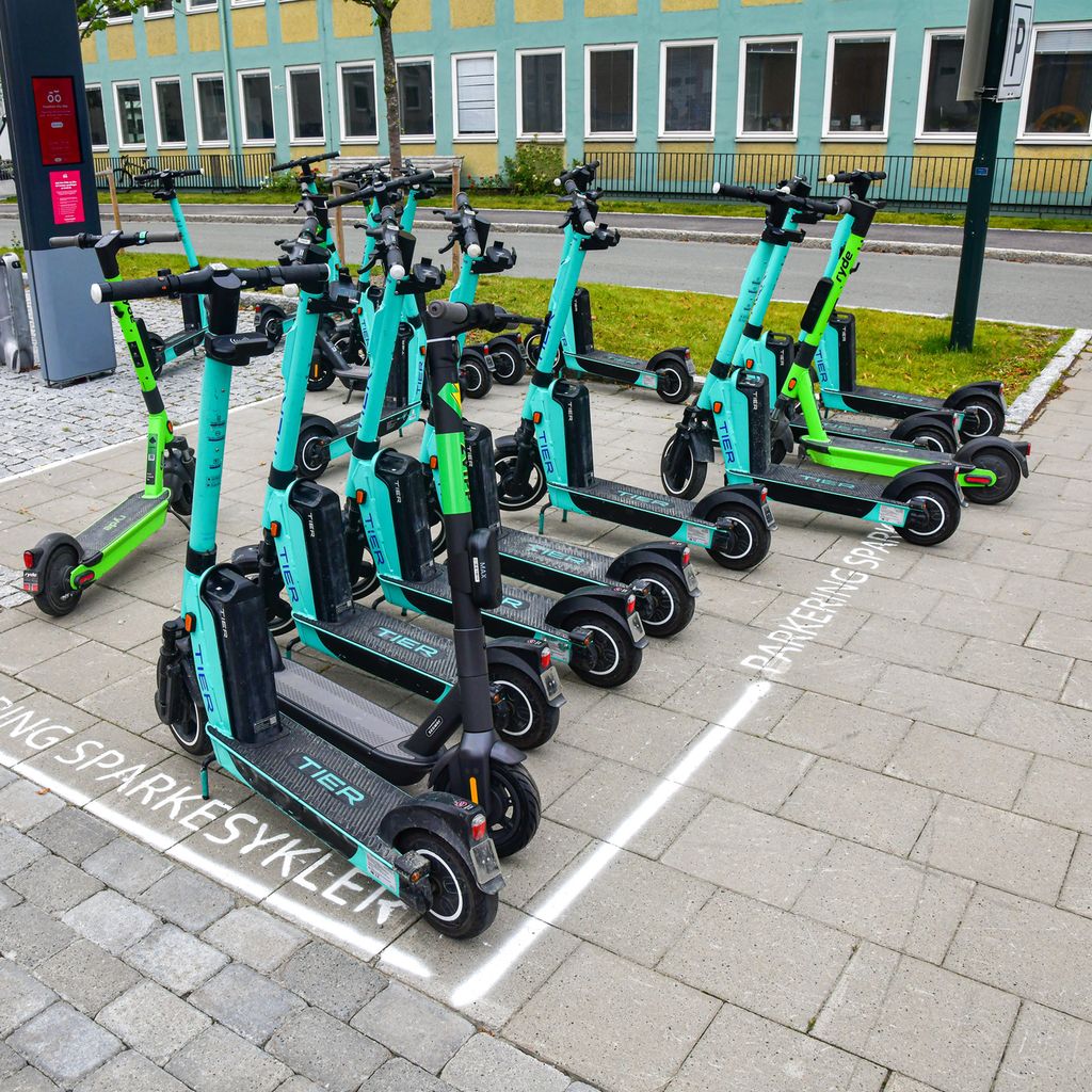Omtrent 15 sparkesykler fra forskjellige utleiefirmaer. Sparkesyklene er parkert delvis inni et rektangel merket opp med hvit maling på bakken. Inni rektangelet står det skrevet "Parkering sparkesykler". Foto.
